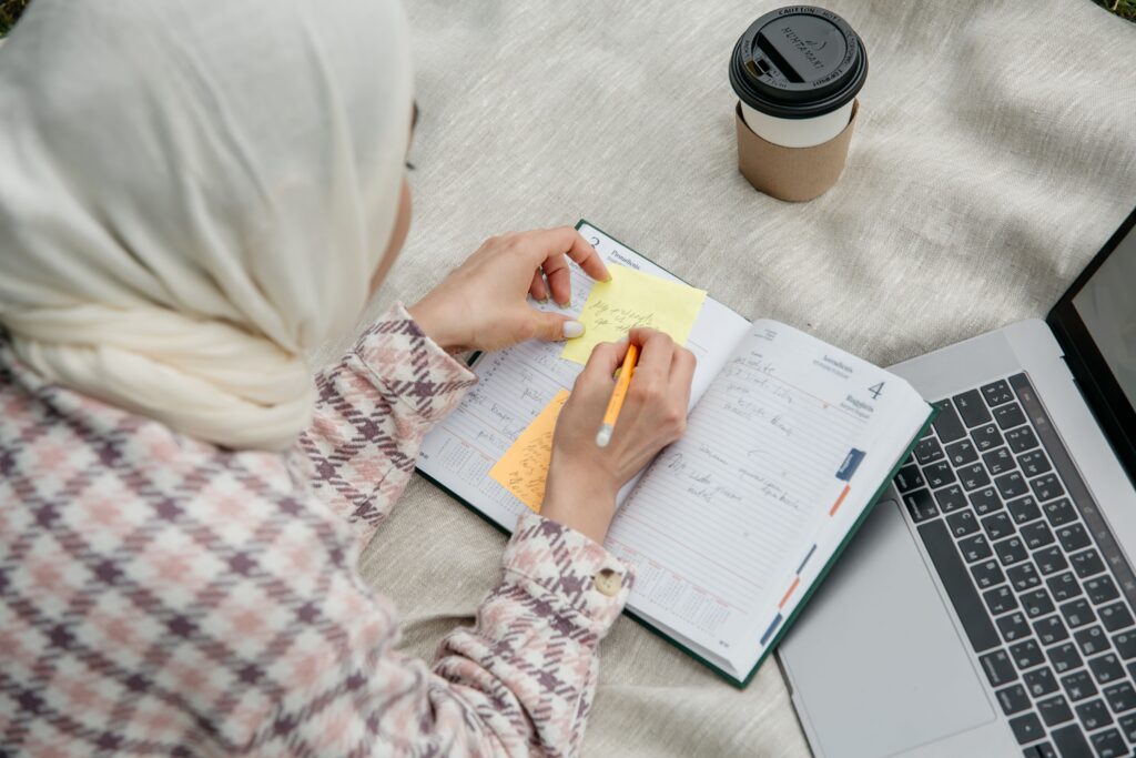Woman in hijab writes notes in a planner with laptop and coffee outdoors.